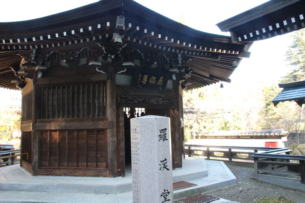 「羅漢堂」と刻まれた石柱が手前に建つ六角形のお堂(羅漢堂)の正面カット