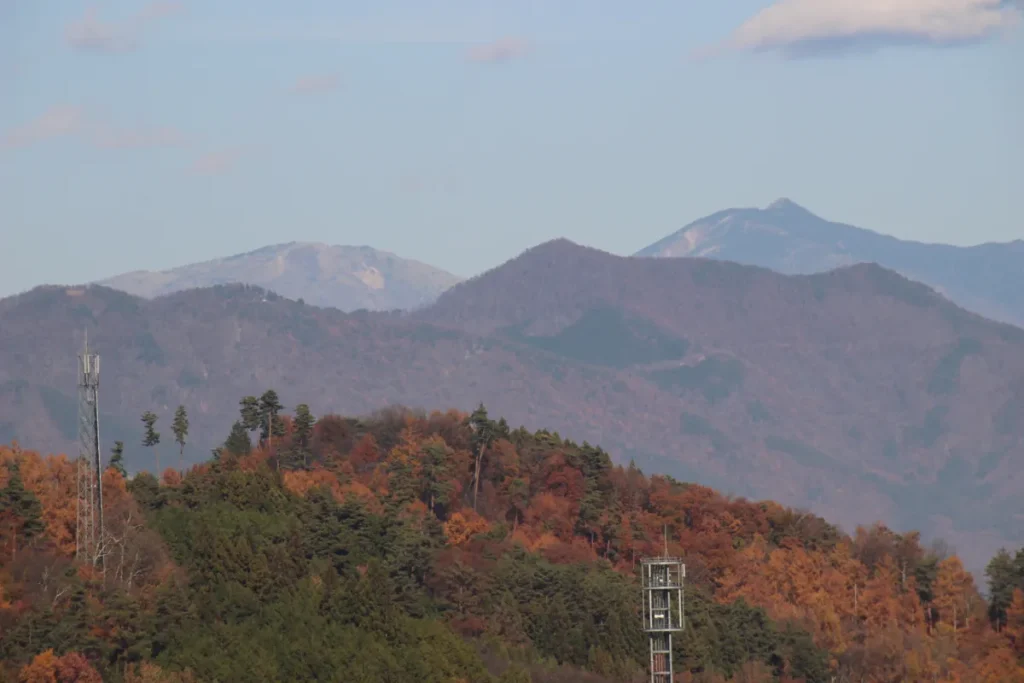 山頂に電波塔が建つ紅葉に染まった里山と、その奥に見える遠方の山脈