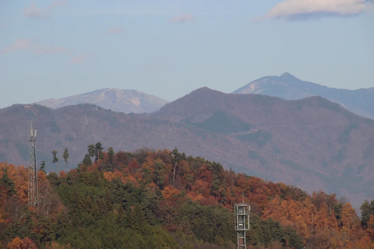 山頂に電波塔が建つ紅葉に染まった里山と、その奥に見える遠方の山脈