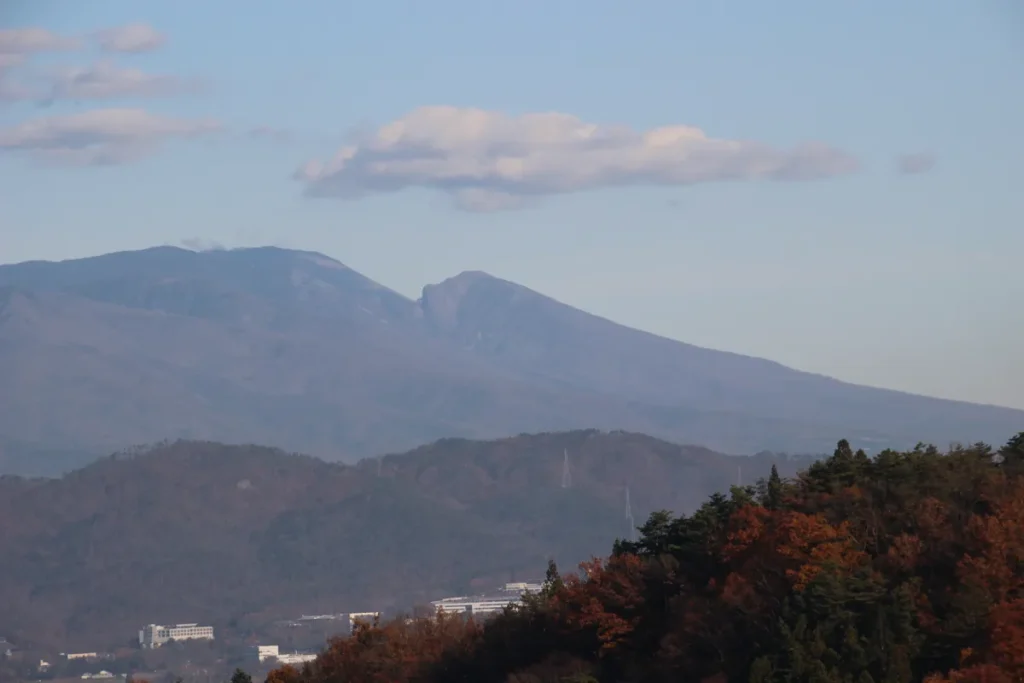手前の美しい紅葉に彩られた里山の斜面と、奥に広がる雄大な信州の山岳パノラマ
