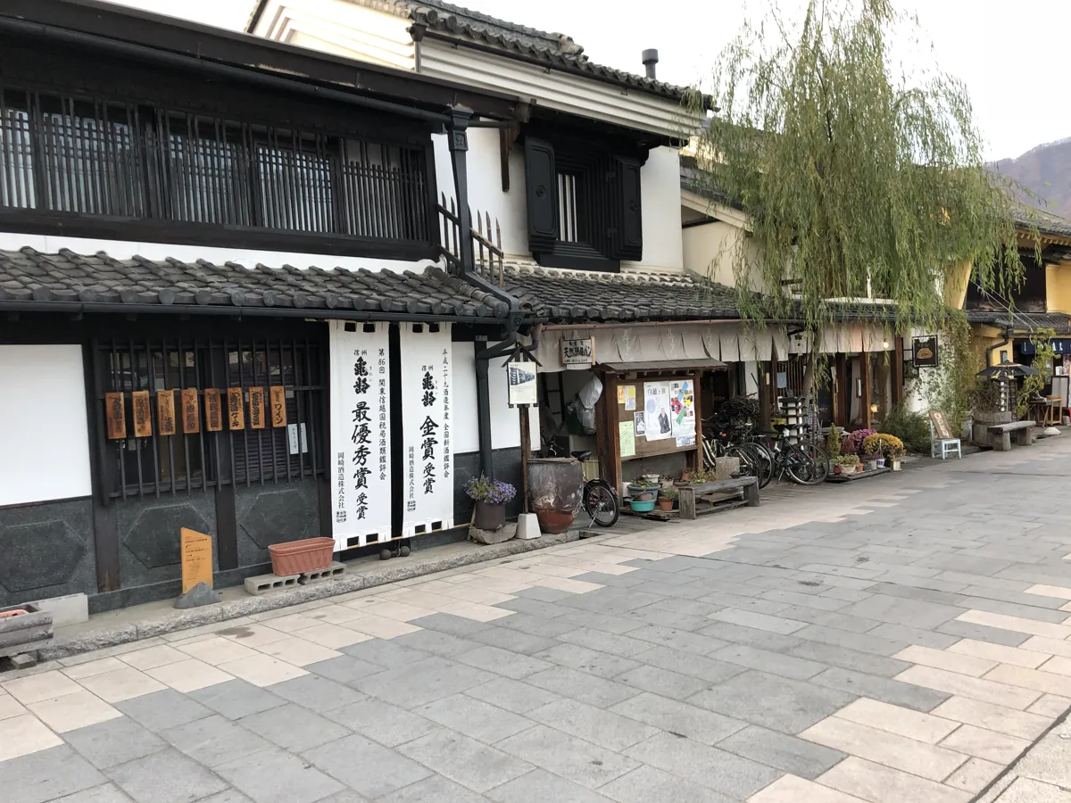 通りの名である柳の緑の枝と、歴史ある岡崎酒造の重厚な蔵造りの建物が調和する美しい通り沿いの光景