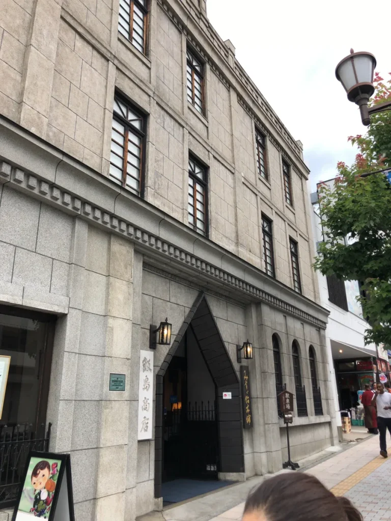 登録有形文化財のプレートが掲げられた飯島商店の洋風建築外観と、店頭に置かれた可愛らしいキャラクター看板